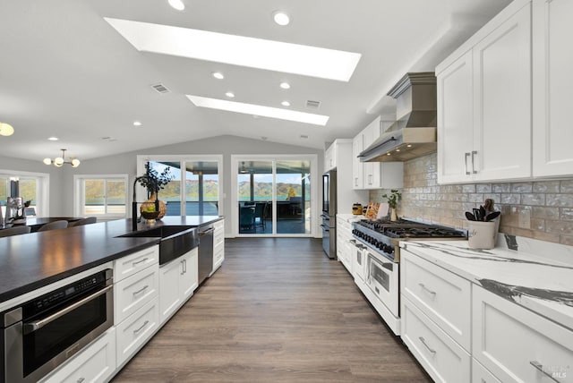 kitchen with wall chimney exhaust hood, appliances with stainless steel finishes, dark hardwood / wood-style floors, vaulted ceiling with skylight, and white cabinets