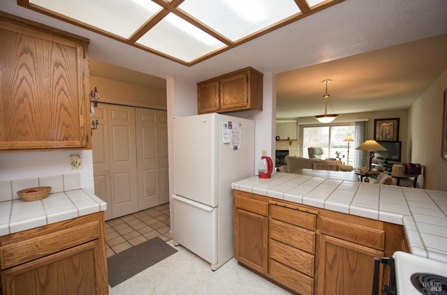 kitchen with open floor plan, tile counters, freestanding refrigerator, brown cabinetry, and a fireplace
