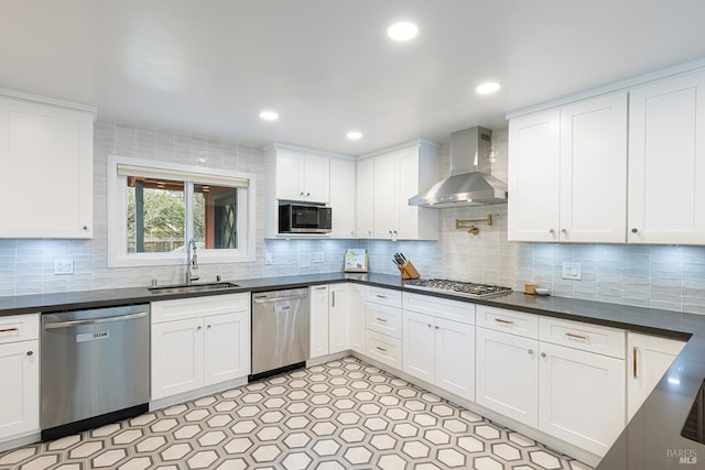 kitchen with white cabinets, dark countertops, appliances with stainless steel finishes, wall chimney range hood, and a sink