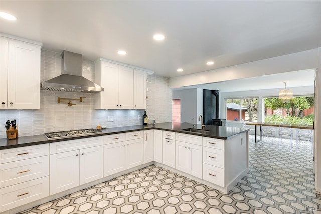 kitchen with dark countertops, a peninsula, wall chimney range hood, stainless steel gas stovetop, and a sink