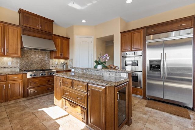 kitchen featuring appliances with stainless steel finishes, a kitchen island, light stone countertops, decorative backsplash, and wall chimney range hood