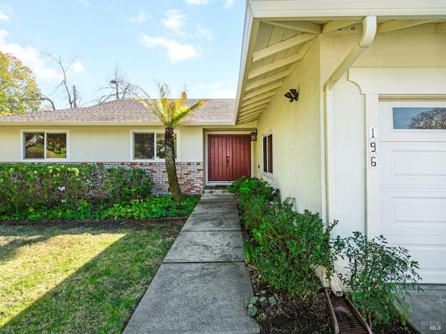 view of exterior entry featuring a yard, brick siding, a shingled roof, and stucco siding