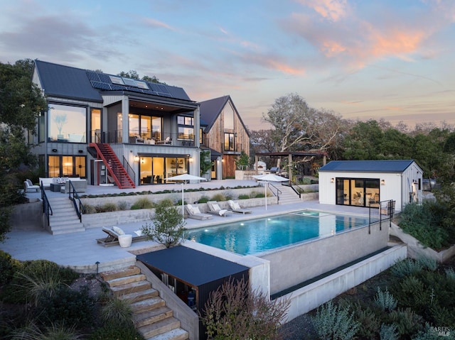 back of house at dusk with an outbuilding, an infinity pool, solar panels, a patio area, and stairs