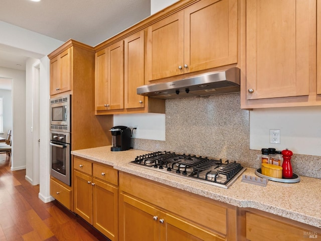 kitchen with light stone counters, dark wood-style floors, stainless steel appliances, decorative backsplash, and under cabinet range hood