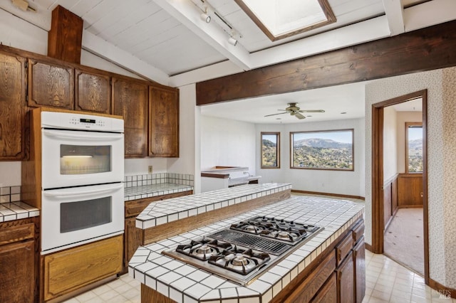 kitchen with white double oven, tile countertops, a center island, stainless steel gas cooktop, and beam ceiling