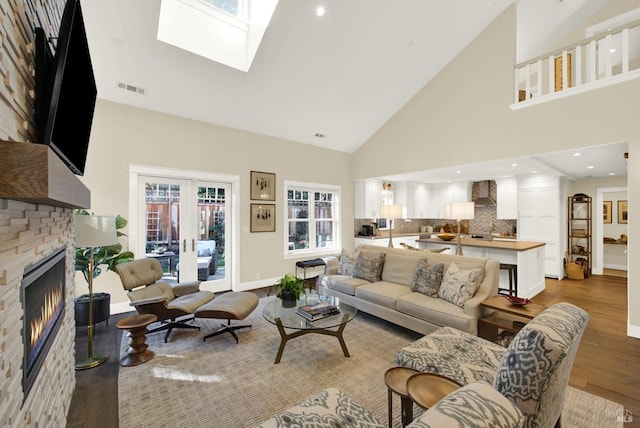 living room featuring high vaulted ceiling, a skylight, visible vents, light wood finished floors, and a glass covered fireplace