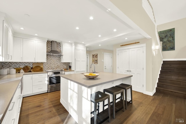 kitchen with electric stove, wall chimney exhaust hood, white cabinetry, an island with sink, and a kitchen bar