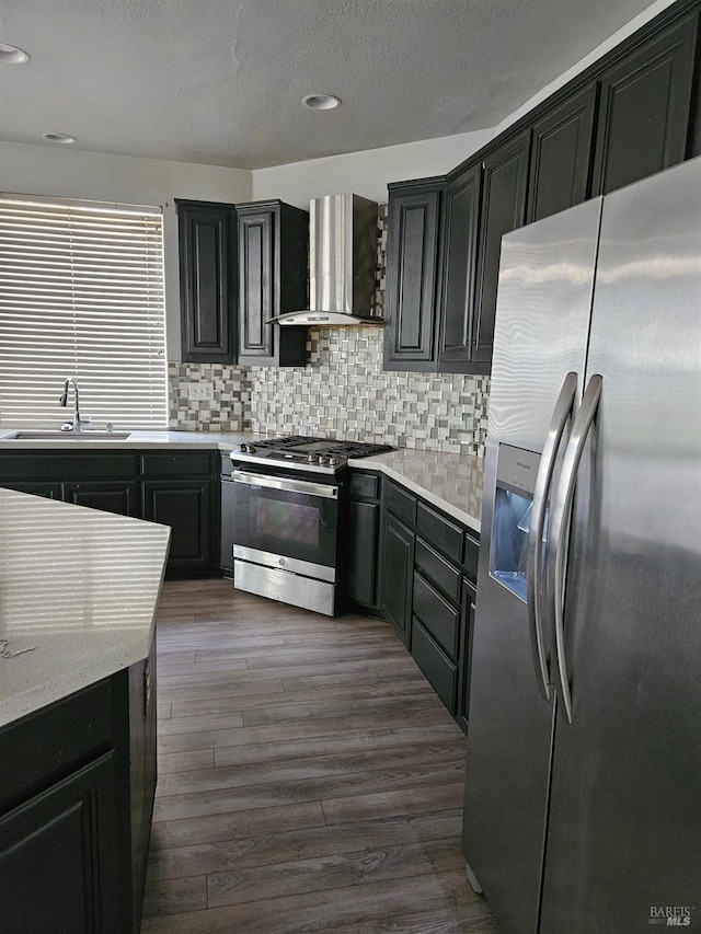 kitchen with stainless steel appliances, dark wood-type flooring, a sink, wall chimney range hood, and dark cabinets