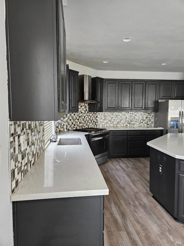 kitchen with dark cabinets, stainless steel appliances, a sink, light countertops, and wall chimney range hood