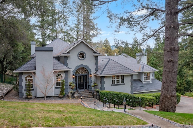 view of front facade with stucco siding, roof with shingles, a chimney, and a front yard