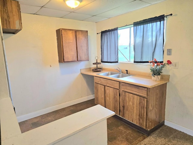 kitchen with brown cabinets, a paneled ceiling, light countertops, a sink, and baseboards