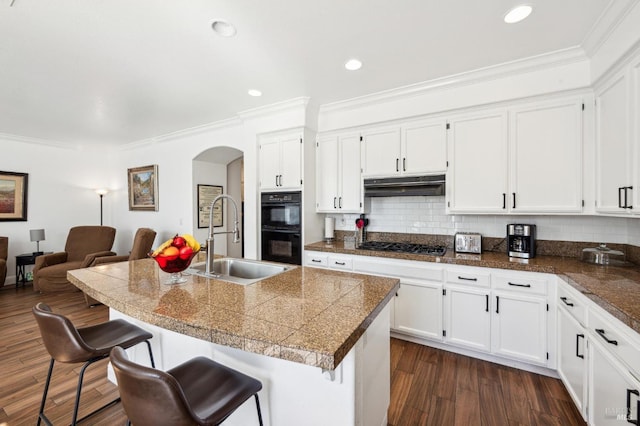 kitchen with dobule oven black, tile counters, arched walkways, under cabinet range hood, and a sink