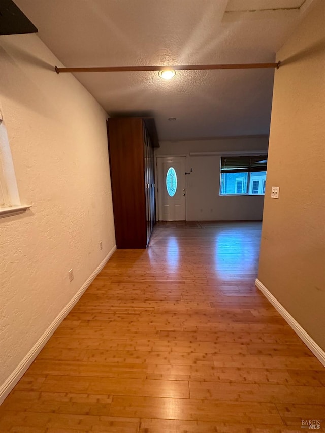 hall with a textured ceiling, a textured wall, light wood-style flooring, and baseboards