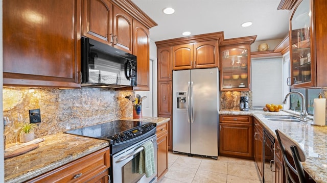 kitchen featuring brown cabinets, appliances with stainless steel finishes, glass insert cabinets, a sink, and light stone countertops