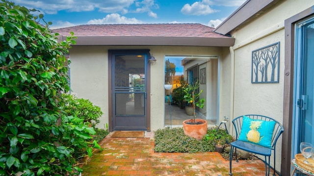 doorway to property with a shingled roof, a patio area, and stucco siding