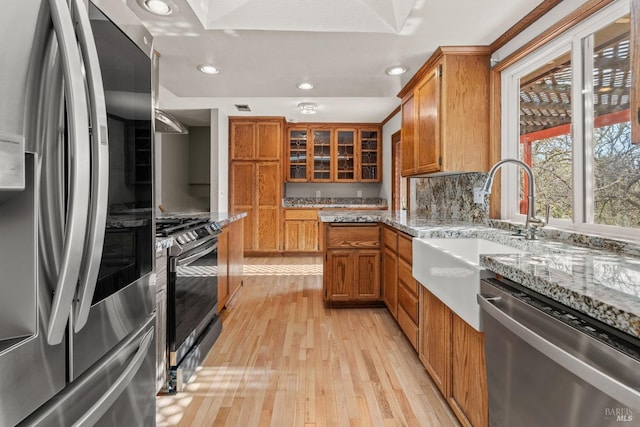 kitchen with light stone counters, stainless steel appliances, brown cabinetry, glass insert cabinets, and a sink