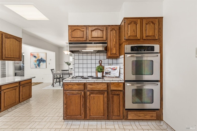 kitchen featuring brown cabinets, tasteful backsplash, white stovetop, stainless steel double oven, and under cabinet range hood