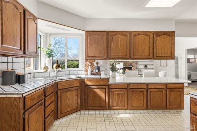 kitchen featuring tile countertops, decorative backsplash, brown cabinetry, a sink, and a peninsula