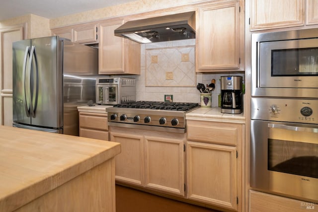 kitchen with wall chimney exhaust hood, tile counters, stainless steel appliances, and light brown cabinetry