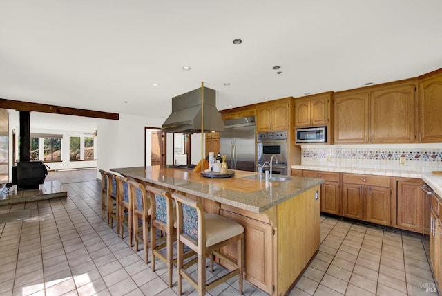 kitchen featuring an island with sink, built in appliances, a wood stove, island exhaust hood, and a kitchen bar