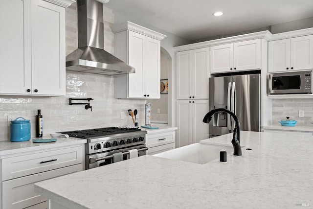 kitchen featuring light stone counters, a sink, white cabinets, wall chimney range hood, and appliances with stainless steel finishes