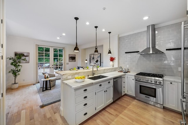 kitchen with light wood-style flooring, stainless steel appliances, a peninsula, a sink, and wall chimney exhaust hood