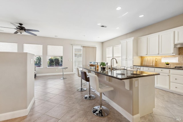 kitchen with visible vents, decorative backsplash, white cabinetry, an island with sink, and dark stone countertops