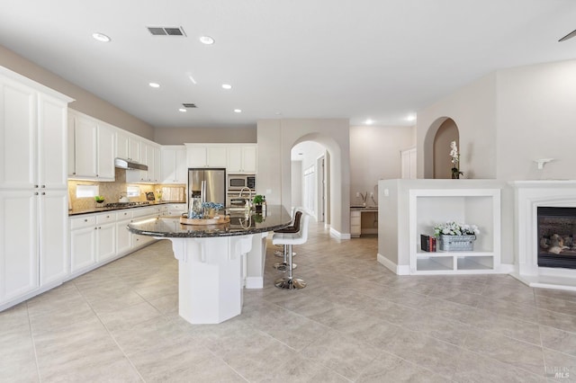 kitchen featuring white cabinets, dark stone counters, appliances with stainless steel finishes, a kitchen island with sink, and a kitchen bar