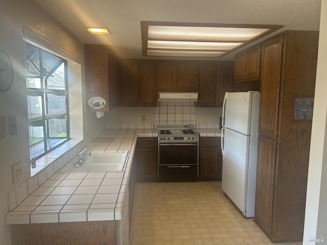 kitchen featuring tile countertops, stainless steel stove, under cabinet range hood, a sink, and freestanding refrigerator