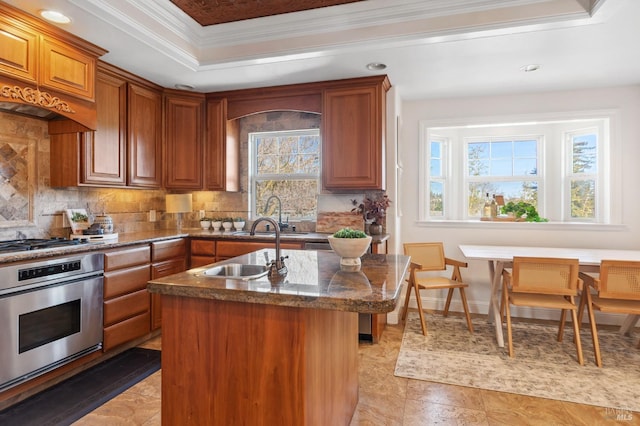 kitchen featuring a center island with sink, stainless steel appliances, a raised ceiling, brown cabinetry, and a sink