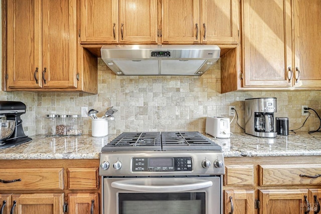 kitchen with light stone counters, backsplash, stainless steel gas stove, and under cabinet range hood