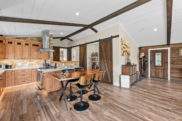 kitchen featuring a barn door, wall chimney exhaust hood, wood finished floors, vaulted ceiling with beams, and stainless steel range with gas cooktop