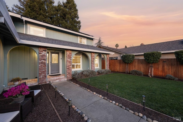 property entrance with roof with shingles, brick siding, a lawn, and fence