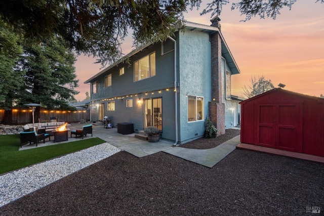 back of house at dusk with a fire pit, a patio, an outbuilding, a shed, and stucco siding