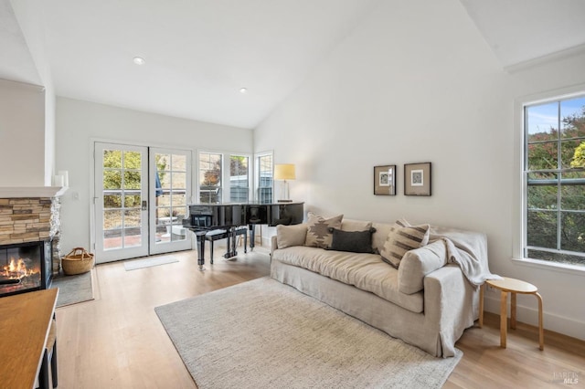 living room featuring light wood finished floors, baseboards, high vaulted ceiling, and a stone fireplace