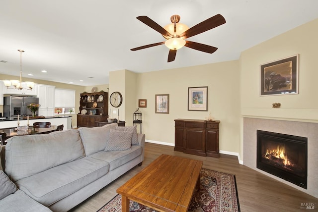 living room featuring ceiling fan with notable chandelier, dark wood-style flooring, a tile fireplace, and baseboards