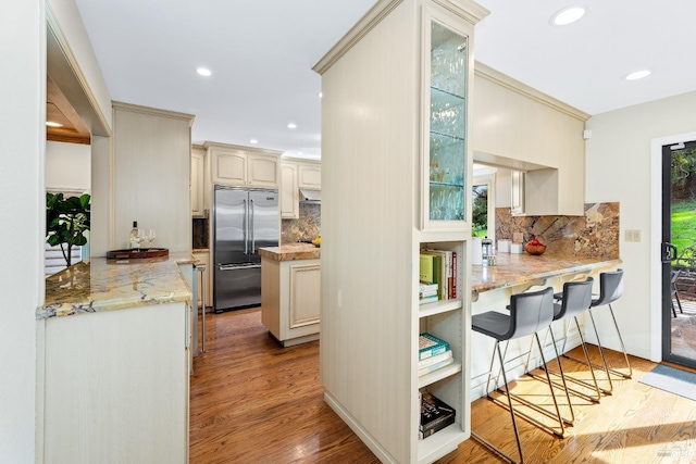 kitchen with glass insert cabinets, a peninsula, light stone countertops, stainless steel built in refrigerator, and a kitchen bar