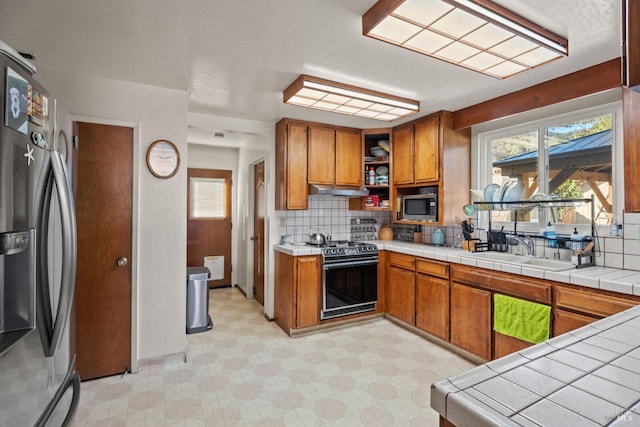 kitchen with tile counters, stainless steel appliances, light floors, open shelves, and a sink