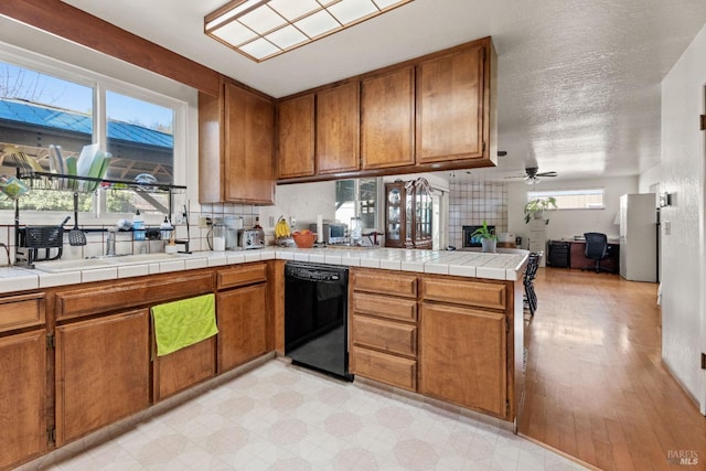 kitchen with black dishwasher, brown cabinetry, tile countertops, freestanding refrigerator, and a peninsula