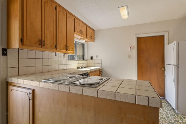 kitchen with a peninsula, brown cabinets, tile counters, freestanding refrigerator, and stainless steel gas stovetop