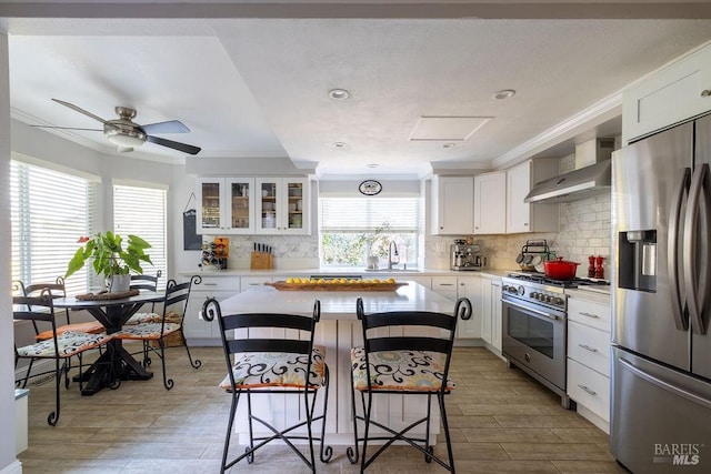 kitchen featuring stainless steel appliances, white cabinets, ornamental molding, wall chimney range hood, and a kitchen bar