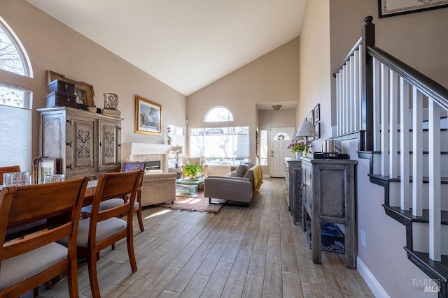 living room featuring baseboards, stairway, light wood-style floors, a fireplace, and high vaulted ceiling