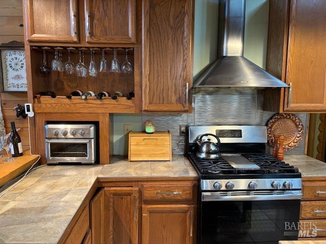 kitchen with tile countertops, decorative backsplash, brown cabinetry, gas range, and wall chimney exhaust hood