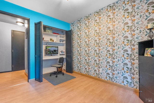 office area featuring wallpapered walls, built in desk, baseboards, and wood finished floors