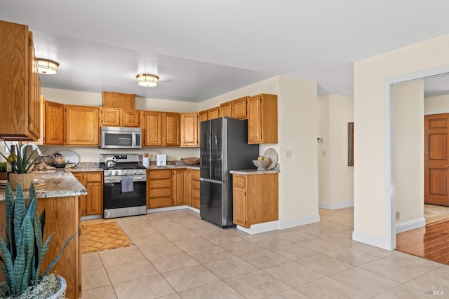 kitchen featuring baseboards, appliances with stainless steel finishes, brown cabinetry, and light stone counters