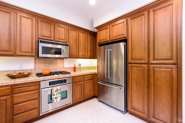 kitchen featuring tasteful backsplash, brown cabinetry, tile countertops, stainless steel appliances, and light floors