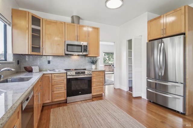 kitchen featuring light stone counters, light brown cabinets, stainless steel appliances, a sink, and glass insert cabinets