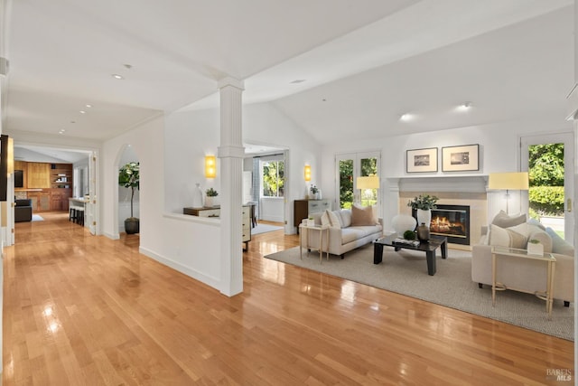 living area with lofted ceiling, baseboards, light wood finished floors, a glass covered fireplace, and decorative columns