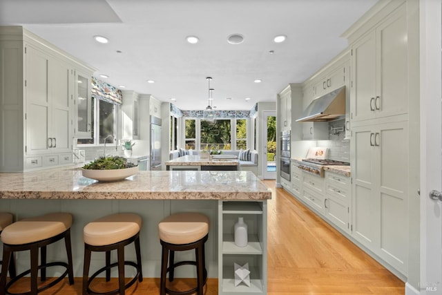 kitchen featuring light wood finished floors, glass insert cabinets, decorative light fixtures, a peninsula, and under cabinet range hood