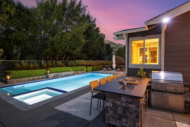 pool at dusk with exterior bar, an in ground hot tub, a fenced in pool, and area for grilling